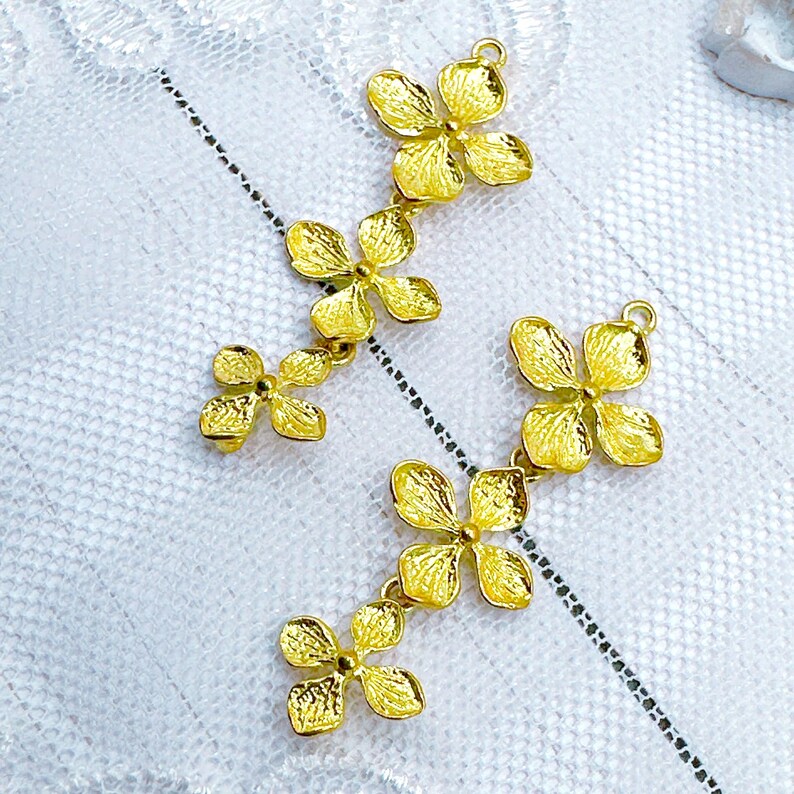 May include: Two gold-tone flower charms with a dangling design. Each charm features four flower petals and a loop at the top for attaching to a necklace or bracelet.
