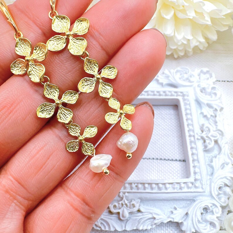 May include: Gold flower-shaped earrings with a dangling pearl accent. The earrings are made of a delicate metal and have a floral design.