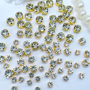 May include: A collection of sparkling, clear rhinestones with gold-colored metal settings. The rhinestones vary in size and are scattered across a white surface. Some are round, and others are square-shaped.