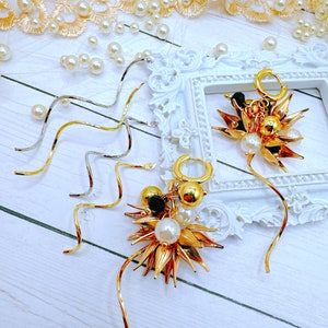 May include: Two gold dangle earrings with black and white beads and gold leaf accents. The earrings are hanging from a white frame.