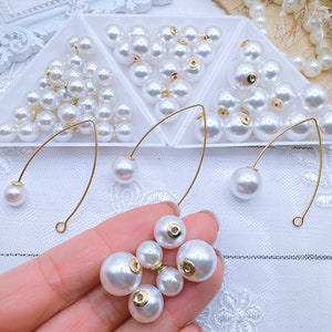 May include: A close-up of white pearl beads in three triangular containers. The pearls are arranged in a random pattern. There are three gold-colored wire loops with a pearl attached to each end. A hand is holding five white pearls with gold accents.