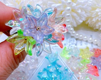 Könnte beinhalten: Ein blütenförmiger Ring mit durchscheinenden Blütenblättern in Blau-, Grün-, Rosa- und Lilatönen. Der Ring hat eine goldfarbene Mitte und ist von anderen blütenförmigen Verzierungen umgeben.