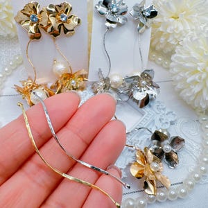 May include: A pair of gold flower earrings with pearl accents and a pair of silver flower earrings with pearl accents. The earrings are displayed on a white surface with a hand holding a gold and silver chain.