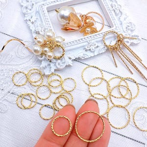 May include: A collection of gold-colored jump rings, some are twisted, some are smooth. The rings are scattered on a white surface. A hand is holding two of the rings.