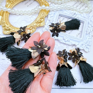 May include: A close-up of a hand holding a pair of black tassel earrings with gold accents. The earrings feature black flower-shaped charms with gold centers. The earrings are on a white background.
