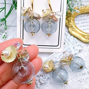 May include: A pair of earrings with a clear glass sphere and gold accents. The earrings are adorned with small white pearls, iridescent beads, and gold leaf accents. The earrings are hanging from a gold hook.