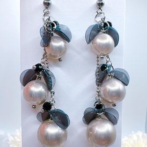 May include: A pair of long, dangling earrings with multiple pearl-like beads and translucent teal-colored leaves. The earrings are silver-toned and have a delicate chain.