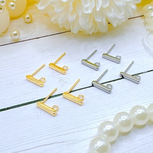 May include: A set of earring findings in gold and silver. The findings are bar-shaped with a loop at one end and a post at the other. The findings are arranged on a white wooden surface.