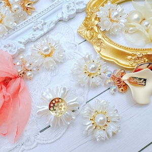 May include: A collection of white pearl flower embellishments with gold accents. The embellishments are arranged on a white lace surface. The embellishments are perfect for adding a touch of elegance to any craft project.