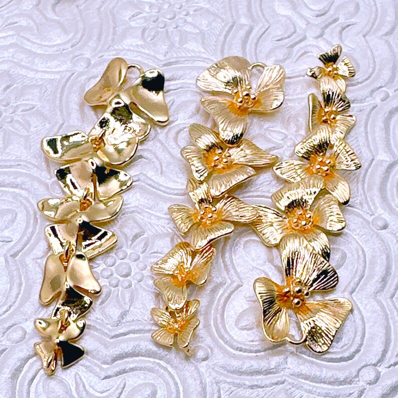 May include: Two strands of gold-tone metal flower charms. Each strand features a series of small, delicate flower charms connected together. The charms are in the shape of a simple flower with five petals.