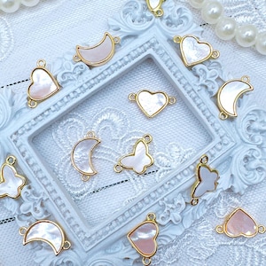 May include: Assortment of white and pink shell charms with gold accents. The charms are shaped like hearts, moons, butterflies, and a simple oval. The charms are displayed on a white background with a gold frame.