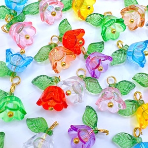 May include: A collection of colorful, translucent flower charms in various shades including pink, red, orange, yellow, green, blue, and purple. Each charm features a gold-colored loop and a small gold bead in the center. Green leaf accents are also present.