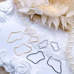 May include: An assortment of abstract-shaped metal jewelry components in gold, silver, and black. The components are arranged on a white surface with floral and pearl accents, suggesting a craft or jewelry-making theme.