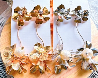 Op de afbeelding: Twee paar oorhangers met bloemvormige bedels. Het ene paar is goud met heldere strass steentjes en het andere is zilver met heldere strass steentjes.