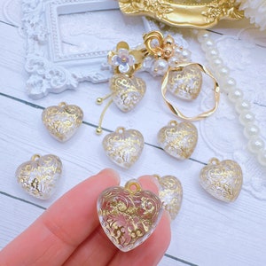 May include: Eight gold heart-shaped charms with a white background and intricate gold designs. The charms have a loop at the top for attaching to jewelry or other crafts.
