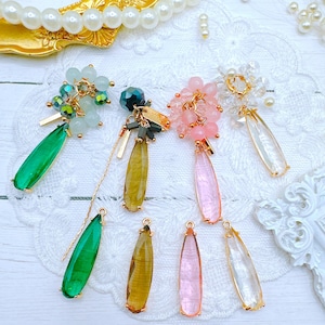 May include: Assortment of teardrop-shaped glass beads with gold accents. The beads are in various colors, including green, brown, pink, and clear. The beads are arranged on a white surface with a lace pattern.