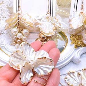 May include: A pair of white pearl-like flower earrings with gold accents and a gold chain. The earrings are hanging from a white surface.