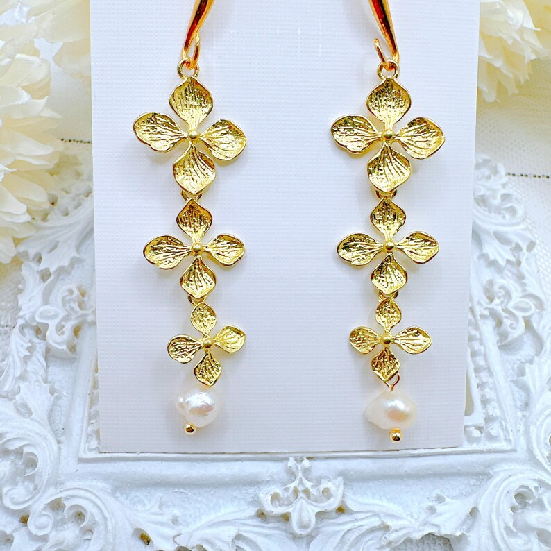 May include: Gold dangle earrings with three flower-shaped charms and a pearl at the bottom.