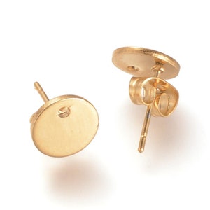 May include: Two gold-colored stud earrings with a flat, circular design. One earring is shown with the post and back, while the other displays the front. The earrings are set against a white background.