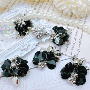 May include: An assortment of black sequin and silver-toned jewelry pieces. The set includes earrings, a ring, and a necklace pendant, all featuring a floral design with pearl accents. The pieces are arranged on a white surface.