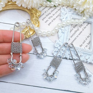 May include: Close-up of three silver safety pin earrings with a woven metal design and dangling clear crystal beads. The earrings are displayed on a white surface with decorative elements in the background.
