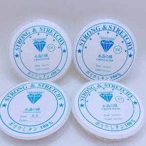May include: Four spools of clear elastic string labeled "Strong & Stretchy" with the text "Crystal String" and "Polyurethane 100%". The spools are labeled with different sizes: 0.4, 0.5, 0.6, and 0.7.