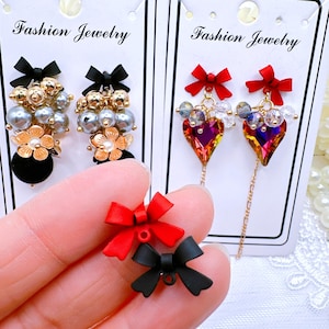 May include: Two pairs of earrings with different styles. One pair features a black bow with a cluster of gray pearls and a gold flower charm. The other pair features a red bow with a heart-shaped crystal charm and a chain.
