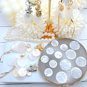 May include: A collection of white mother of pearl shell circles with holes in the center, displayed on a gray plate. The shells are arranged in a circular pattern. There are also several silver and gold charms with beads and shells.
