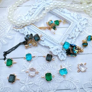 May include: Assortment of green and blue glass and crystal beads and charms with gold accents. The beads are in various shapes, including square, round, and teardrop. The charms are in the shape of flowers and leaves. The beads and charms are arranged on a white lace background.