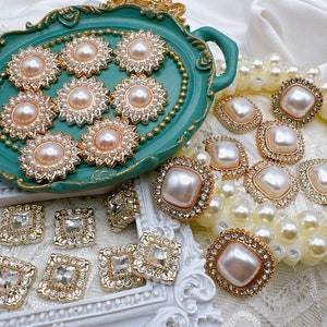 May include: Assortment of pearl and rhinestone embellishments in gold and white tones. The embellishments are arranged on a green tray and a white surface. Some of the embellishments are square shaped and some are round.