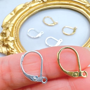 May include: Close-up of silver and gold-colored earring clasps. The clasps are in various sizes and shapes, with a lever-back design. The background includes a gold-colored ornate frame and other earring components.