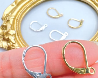 Op de afbeelding: Close-up van zilver- en goudkleurige oorbel sluitingen. De sluitingen zijn in verschillende maten en vormen, met een beugel-back design. De achtergrond bevat een goudkleurig sierlijk frame en andere oorbel componenten.