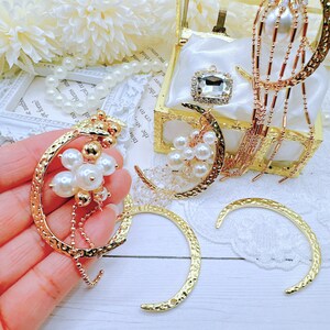 May include: Gold crescent moon earrings with a textured finish. The earrings have a dangling chain with pearl accents. The earrings are displayed on a white surface.
