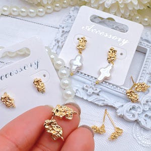 May include: Gold-tone earring findings with a textured, irregular shape. The findings are displayed on white cards with the word "Accessory" printed on them. Some of the findings are paired with white pearl cross charms.
