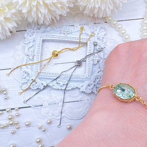 May include: Three gold adjustable bracelets with different charms. One bracelet has a round charm, one has a square charm, and one has a light green oval charm. The bracelets are displayed on a white surface with white pearls.