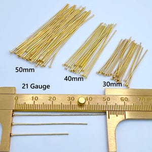 May include: Gold-colored head pins in three sizes: 50mm, 40mm, and 30mm. The image also shows a 21 gauge measurement. These pins are used for jewelry making and crafting. A brass caliper is in the foreground.