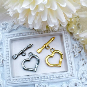 May include: Three metal clasps in the shape of a heart and an arrow. The heart clasps are gold and silver, and the arrow clasp is gold.