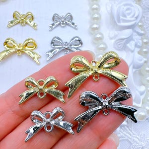 May include: Close-up of several small, metallic bow charms in gold and silver. The charms have a detailed, textured design and a small loop for attaching to jewelry or crafts. The bows are arranged on a light background.