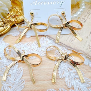 May include: Four pairs of gold-toned bow earrings. The bows are made of a woven metal ribbon. The earrings are on a white background with a lace overlay.