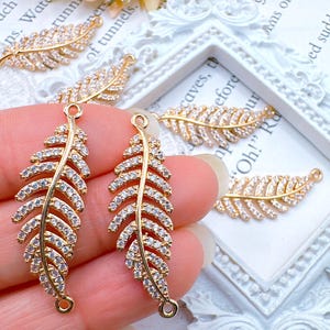 May include: Gold-colored feather-shaped charms with clear, sparkling crystals. The charms have a loop at the top and bottom for attaching to jewelry. Several charms are visible, some held in a hand, others on a white surface.