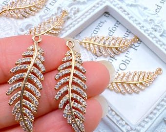 Könnte beinhalten: Goldfarbene, federförmige Charms mit klaren, funkelnden Kristallen. Die Charms haben oben und unten eine Schlaufe zum Befestigen an Schmuck. Mehrere Charms sind sichtbar, einige werden in einer Hand gehalten, andere auf einer weißen Oberfläche.