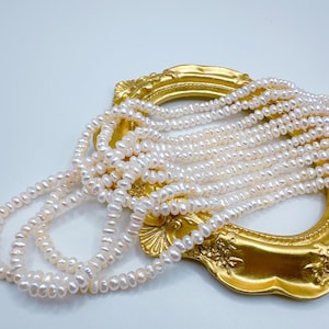 May include: Multiple strands of white freshwater pearls are draped over a gold-colored decorative frame. The pearls have a natural, irregular shape and a soft luster. The frame has an ornate, vintage design. The background is a clean, white surface.