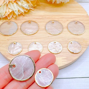 May include: Assortment of white mother of pearl shell circle pendants with gold trim and a hole for stringing. The pendants are arranged on a wooden surface.