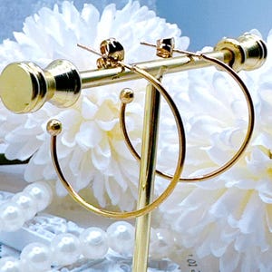 May include: Gold-colored hoop earrings displayed on a gold earring stand. The earrings have a simple, elegant design with a small ball accent on the hoop. The stand also holds a pair of stud earrings. The background features white flowers and a string of pearls.