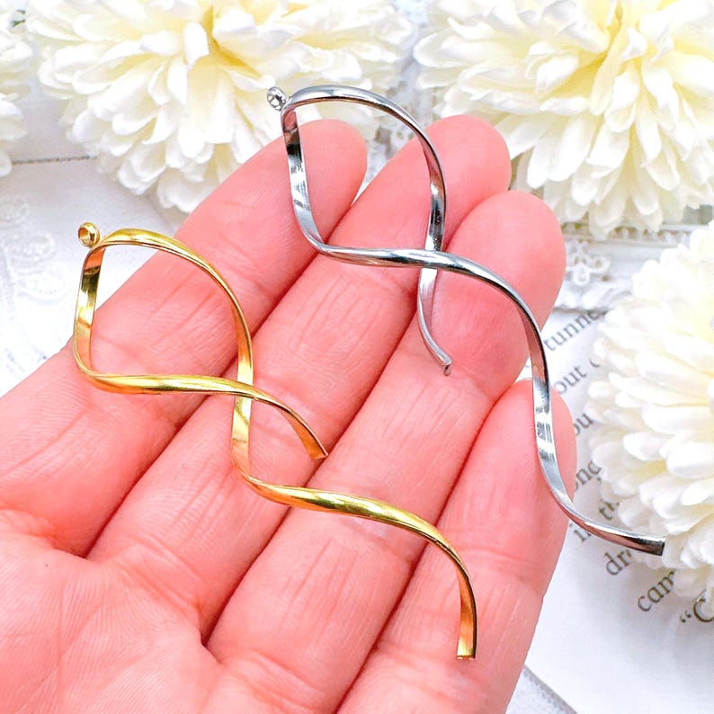 Op de afbeelding: Twee metalen spiraalvormige hangers, &eacute;&eacute;n goudkleurig en &eacute;&eacute;n zilverkleurig, worden in een hand gehouden. De hangers hebben een gedraaid, lintachtig ontwerp met een kleine lus aan de bovenkant om op te hangen. De achtergrond heeft witte bloemen.