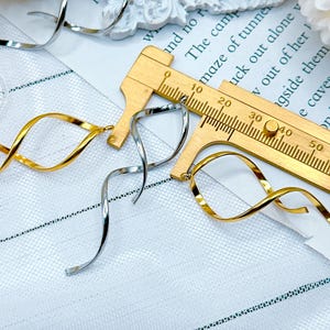 Op de afbeelding: Close-up van goudkleurige en zilverkleurige spiraalvormige oorbellen. De oorbellen zijn gemaakt van dun, gebogen metaal. Op de achtergrond een houten liniaal, met gedeeltelijk zichtbare tekst. De oorbellen liggen op een witte, getextureerde ondergrond.