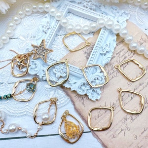 May include: A collection of gold-toned metal earring blanks with various shapes, including teardrop, square, and oval. Some blanks feature a wavy edge. The blanks are arranged on a white surface with a pearl necklace and a piece of paper with text.