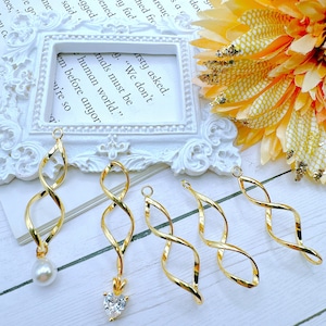 May include: Five gold-colored twisted wire charms with different embellishments. The charms are on a white surface with a white picture frame and a yellow flower in the background. The charms have a pearl, a heart-shaped rhinestone, and three are plain.