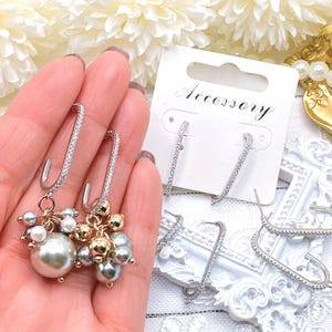 May include: A pair of silver-toned earrings with a row of clear crystals and a cluster of pearls and gold beads. Another pair of earrings with a row of clear crystals is displayed on a white card that says "Accessory".