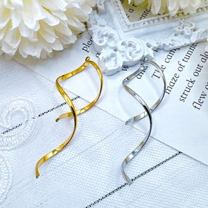 May include: Gold and silver-colored spiral earrings. The earrings have a curved, elongated design, with a loop at the top for attachment. The earrings are displayed on a white surface with a floral accent and text in the background.